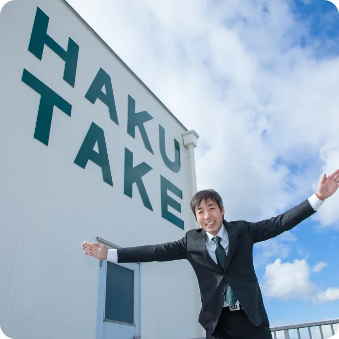 「HAKUTAKE」と大きく書かれた建物の前で、青空の下、両手を広げて笑顔を見せる亀山晋次社長