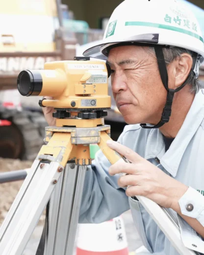 建設現場で、真剣な表情で測量機器を覗き込むT・Iさん。