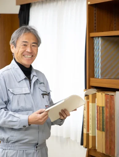 オフィスの本棚の前で、開いた本を両手で持ち、笑顔を浮かべるT・Iさん。